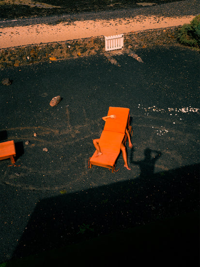 On The Short Romantic Story Between a Chair and a Shadow poster, an individual reclines on an orange beach chair on dark gravel, hand over their head. A second chair is nearby, with the photographers shadows casting across the ground.
