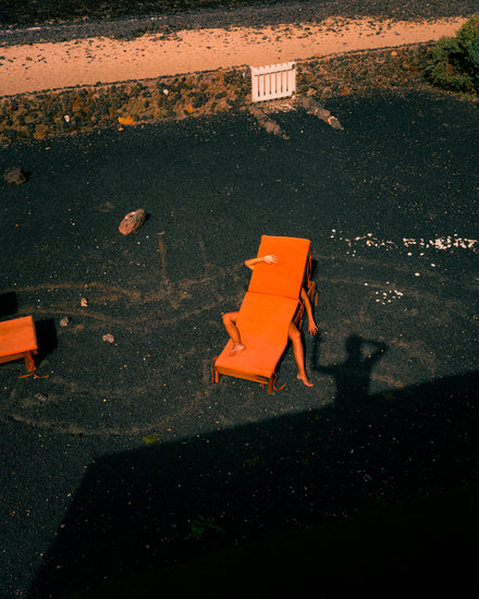 A person is relaxing on two orange chairs, their hand over their face and one leg bent, as intertwined shadows fall on dark gravel. The photographers shadow reaches across the scene with a low stone wall and small fence in the background under a bright sky in The Short Romantic Story Between a Chair and a Shadow poster.