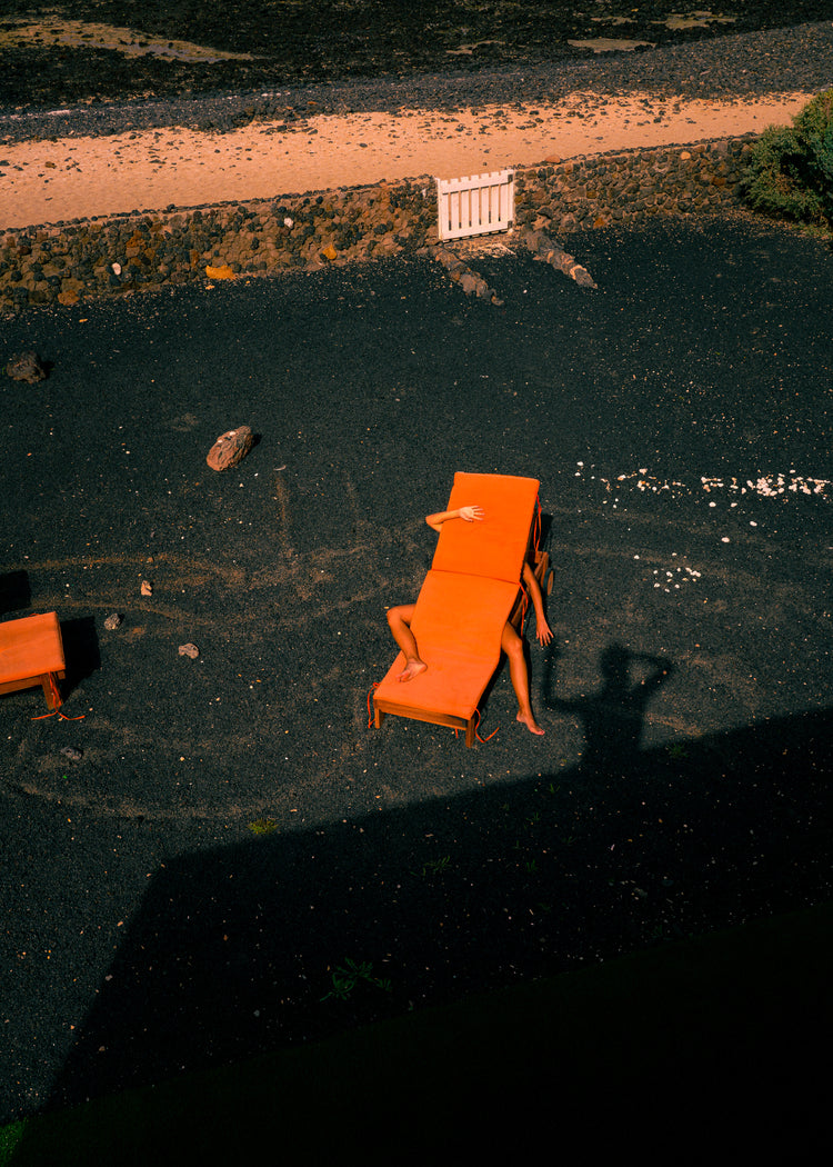 A person is partially hidden under an orange outdoor table and chair set on volcanic rock, with visible arms and legs. Shadows of the photographer create an abstract scene, as depicted in The Short Romantic Story Between a Chair and a Shadow Poster.
