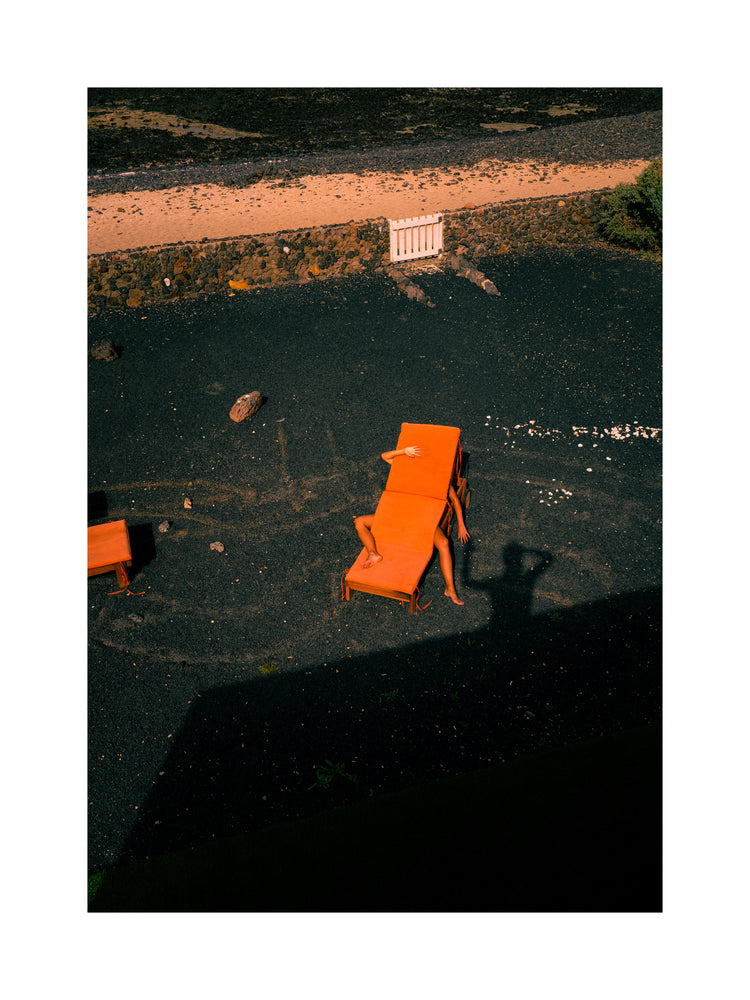 The Short Romantic Story Between a Chair and a Shadow Poster depicts a serene scene of someone reclining on an orange lounge chair amid dark gravel, with shadows artfully cast in the background. A shadow in the foreground shows someone capturing this tranquil moment, framed by a silent boundary wall.