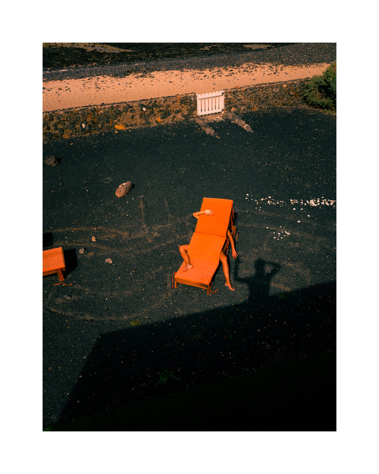 Aerial scene of a person on an orange chair over dark gravel, partially hidden beneath another chair. Their shadowed arm stretches out as the silhouette of someone with raised arms looms nearby. Inspired by The Short Romantic Story Between a Chair and a Shadow poster.