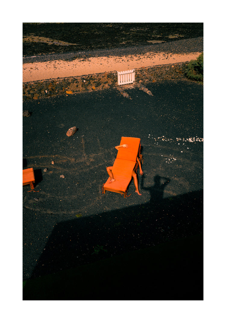 A person relaxes on an orange sunbed, partially shaded, on a gravel surface surrounded by stones like beach pebbles. A wall frames the scene with the photographers shadow subtly captured on the ground, reminiscent of The Short Romantic Story Between a Chair and a Shadow Poster.