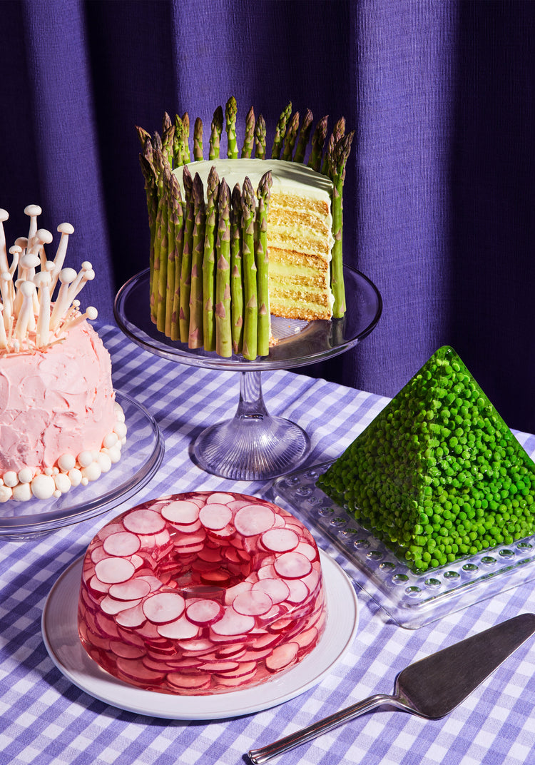 This whimsical still life photograph showcases unconventional cakes adorned with peas, mushrooms, asparagus, and radish on a purple tablecloth, complemented by purple curtains in the background.