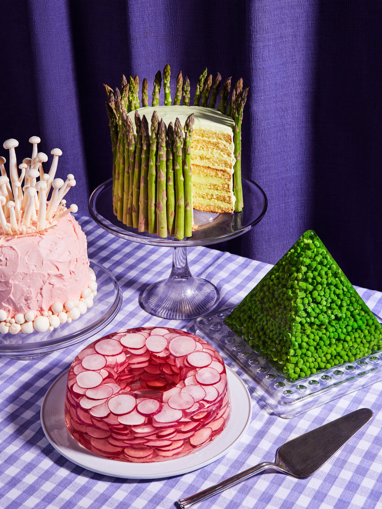 This whimsical still life photograph showcases unconventional cakes adorned with peas, mushrooms, asparagus, and radish on a purple tablecloth, complemented by purple curtains in the background.