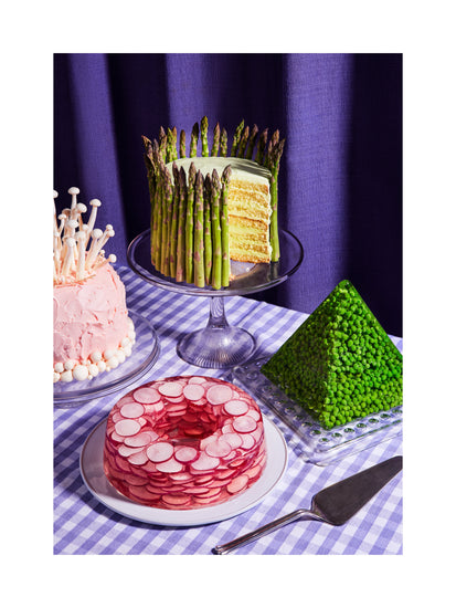 This whimsical still life photograph showcases unconventional cakes adorned with peas, mushrooms, asparagus, and radish on a purple tablecloth, complemented by purple curtains in the background. White border.