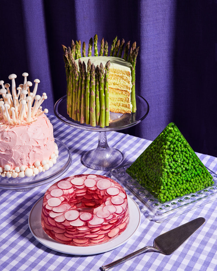 This whimsical still life photograph showcases unconventional cakes adorned with peas, mushrooms, asparagus, and radish on a purple tablecloth, complemented by purple curtains in the background.