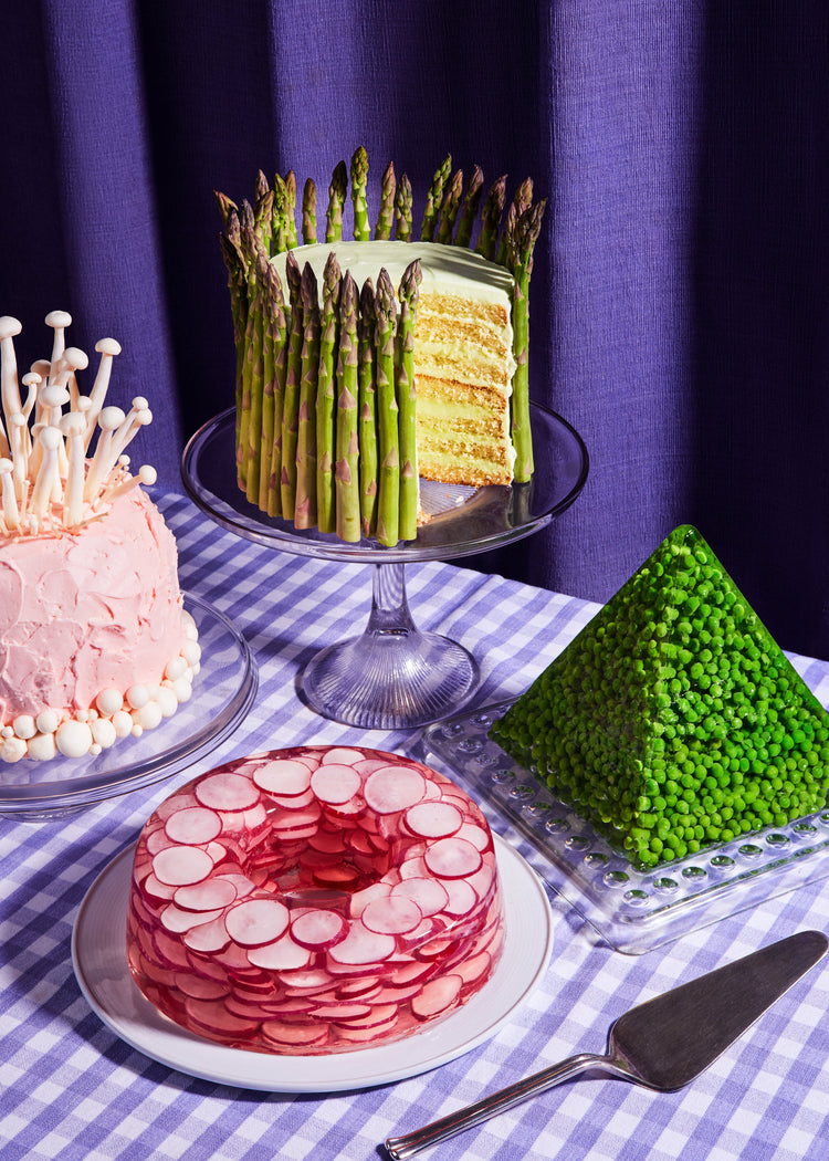 This whimsical still life photograph showcases unconventional cakes adorned with peas, mushrooms, asparagus, and radish on a purple tablecloth, complemented by purple curtains in the background.