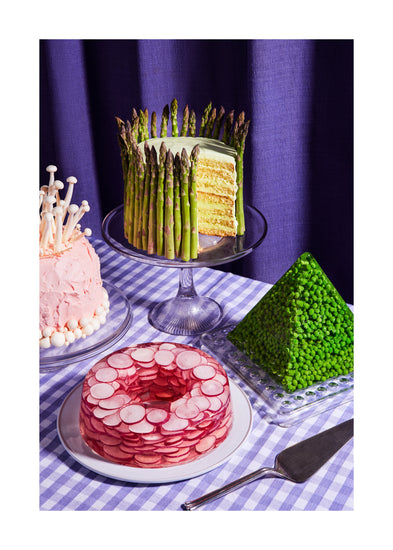 This whimsical still life photograph showcases unconventional cakes adorned with peas, mushrooms, asparagus, and radish on a purple tablecloth, complemented by purple curtains in the background. White border.