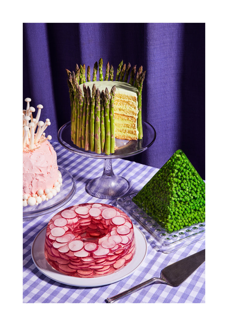 This whimsical still life photograph showcases unconventional cakes adorned with peas, mushrooms, asparagus, and radish on a purple tablecloth, complemented by purple curtains in the background. White border.