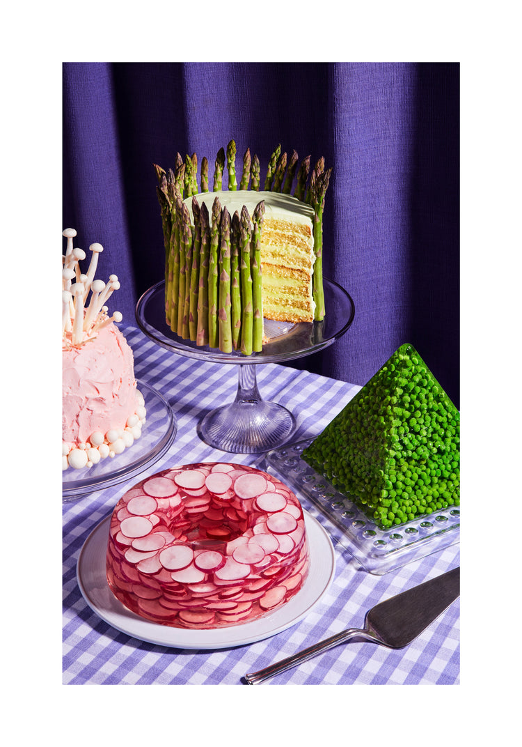 This whimsical still life photograph showcases unconventional cakes adorned with peas, mushrooms, asparagus, and radish on a purple tablecloth, complemented by purple curtains in the background. White border.