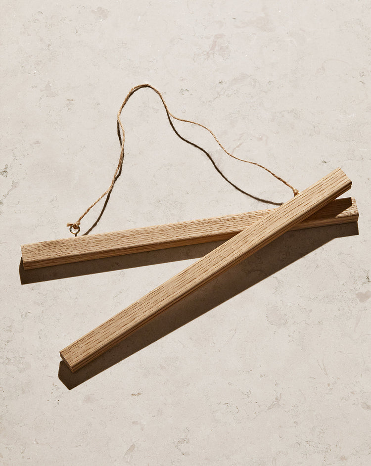 The Poster hanger Oak consists of two solid oak wooden bars with a thin rope connection and discreet magnets to secure your artwork. They lie parallel on a textured, light surface, showcasing a natural wood grain.