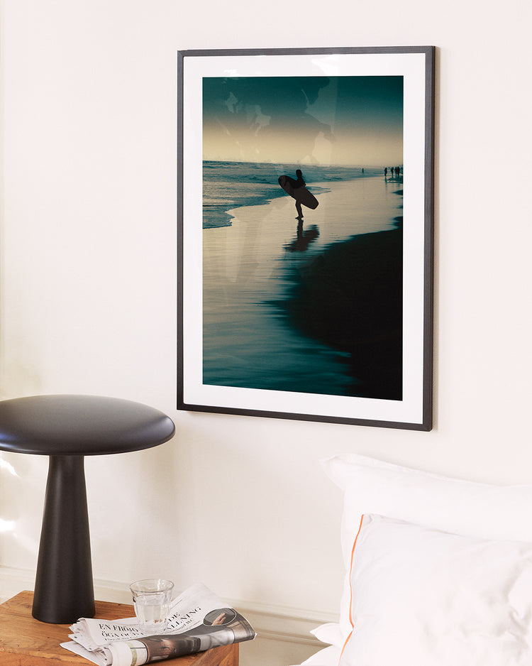 A framed Before Dawn Poster captures a surfers silhouette on dawn patrol, carrying a surfboard at sunset with reflections in the water. Nearby, a black round table holds magazines and a glass, bordered by part of a bed in view.