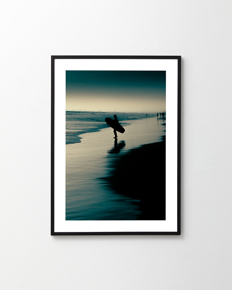 The Before Dawn Poster showcases a framed image of a silhouetted surfer on a dawn patrol, strolling along the beach at sunset. The shoreline reflects the deep blue and teal sky and water, creating a serene and tranquil setting.