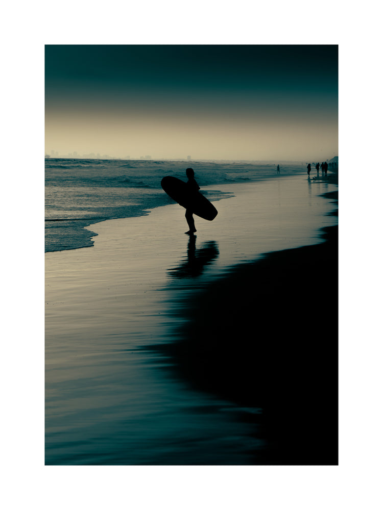 Surfer standing on the shore during low tide under a darkening sky.
