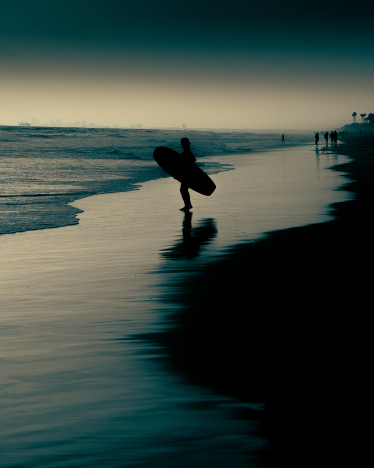 Surfer standing on the shore during low tide under a darkening sky.