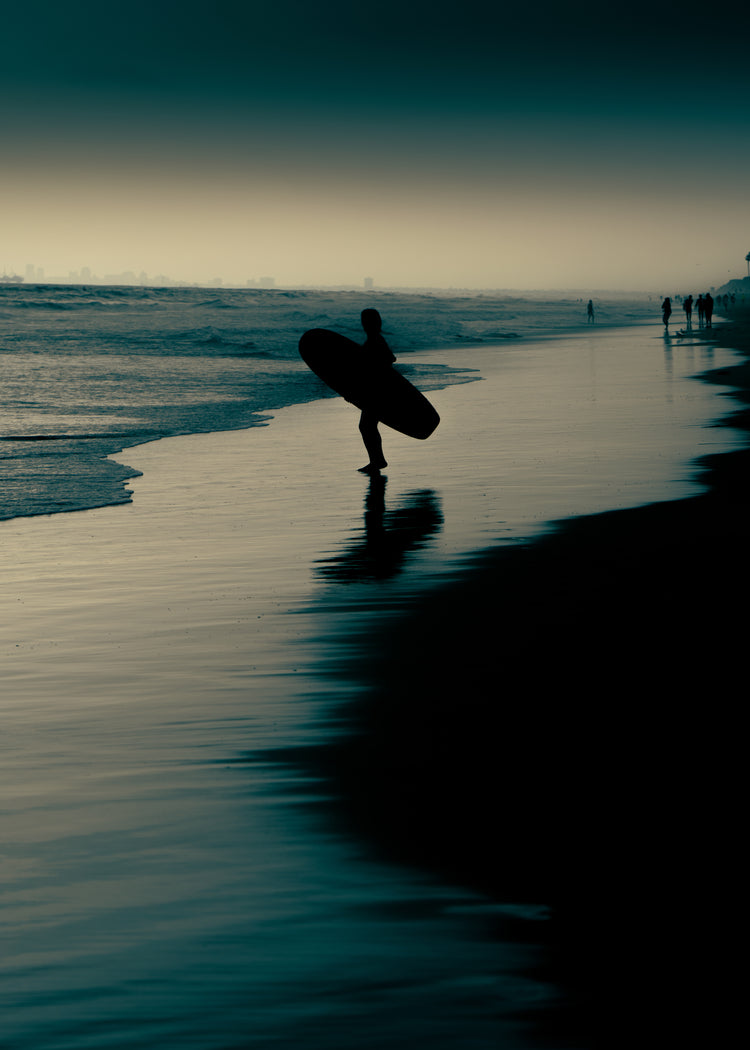 Surfer standing on the shore during low tide under a darkening sky.
