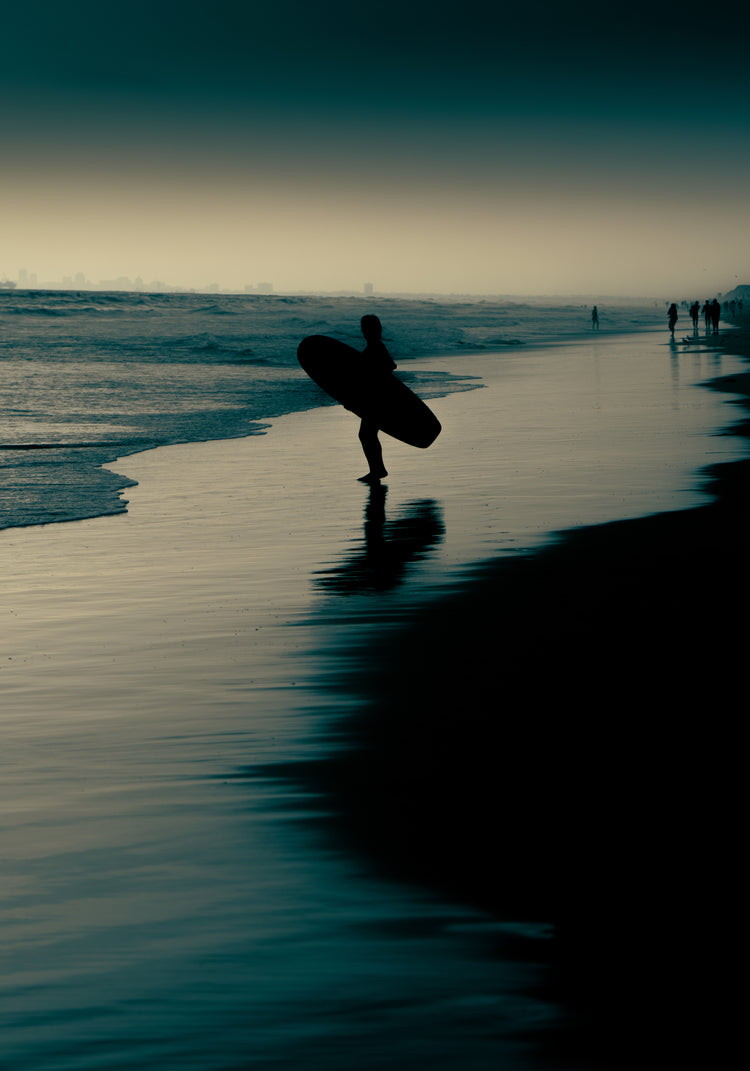 Surfer standing on the shore during low tide under a darkening sky.