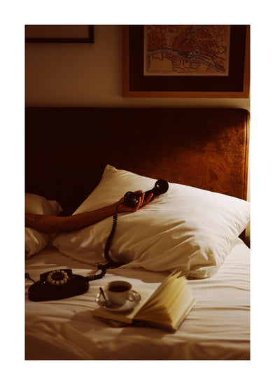 Comfortably nestled in bed while holding a vintage phone handset, a person enjoys the cozy atmosphere with an open book and coffee nearby. Soft lighting and a framed map enhance the retro vibe, captured brilliantly in this Do Not Disturb poster.