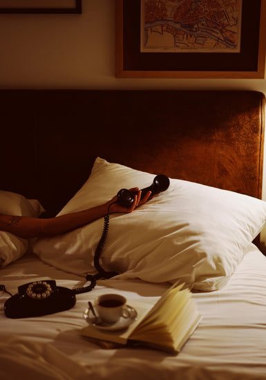A person in bed holds a vintage telephone with its cord trailing, surrounded by white pillows and sheets. Nearby rests an open book and a cup of coffee, creating an ideal scene for the Do Not Disturb poster that captures retro photography or art wall display vibes.