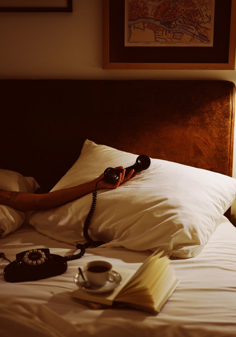 A person in bed holds a vintage telephone with its cord trailing, surrounded by white pillows and sheets. Nearby rests an open book and a cup of coffee, creating an ideal scene for the Do Not Disturb poster that captures retro photography or art wall display vibes.