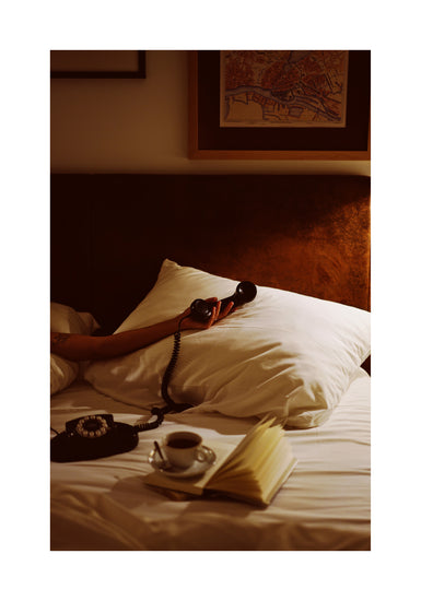 In a dimly lit bedroom, a person lounges on a white bed with an old-school telephone, surrounded by an open book and coffee beside a vintage rotary phone. Above them, the Do Not Disturb Poster art wall and framed map complete the retro scene.