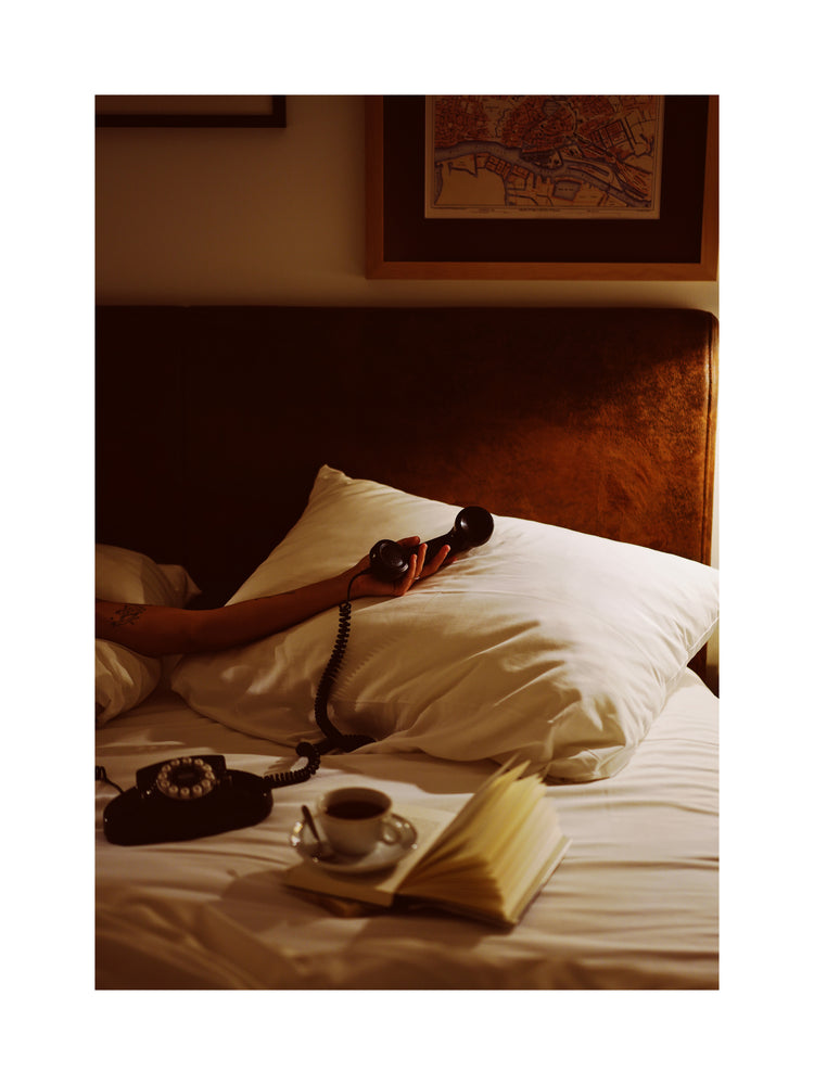 The Do Not Disturb Poster depicts a cozy bedroom with a person in bed holding a vintage phone. On the bed are a pillow, open book, and coffee cup. Above the sofa-like headboard, a map and art pieces add character. Warm lighting completes the inviting scene with retro photography vibes.