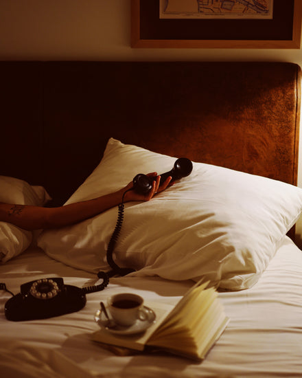 In the Do Not Disturb Poster, a person lies in bed with their arm on a pillow and an old-school telephone in hand. A cup of coffee and an open book sit nearby, while wall artwork and soft lighting create a cozy retro atmosphere.