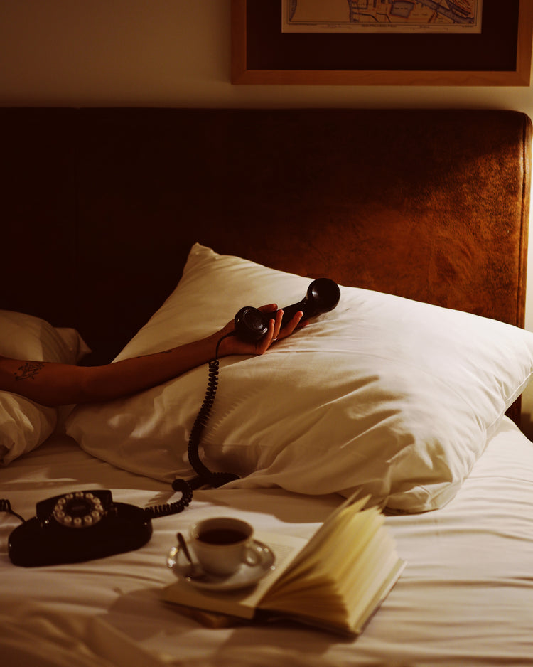 In the Do Not Disturb Poster, a person lies in bed with their arm on a pillow and an old-school telephone in hand. A cup of coffee and an open book sit nearby, while wall artwork and soft lighting create a cozy retro atmosphere.