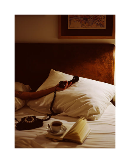 In a dimly lit bedroom, a person holds an old-school telephone on a white pillow. An open book and coffee rest on the bed. The Do Not Disturb Poster, resembling retro photography, hangs above the brown headboard, adding to the cozy nostalgic vibe.