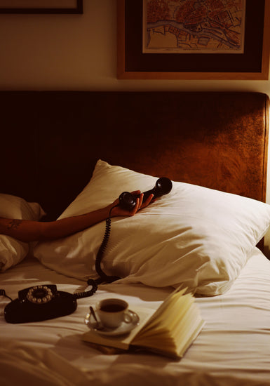 With a Do Not Disturb poster, a person lounges in bed, clutching a rotary phone. A vintage vibe is enhanced by an open book and coffee beside them. The rooms charm is completed by dim lighting and a framed map above the headboard.