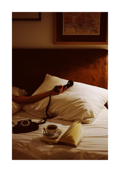 A person lies in bed, arm extended holding a vintage phone receiver. A white pillow and open book share the space, with coffee nearby. The Do Not Disturb Poster featuring a map design hangs above. Warm lighting enhances the cozy, retro vibe.