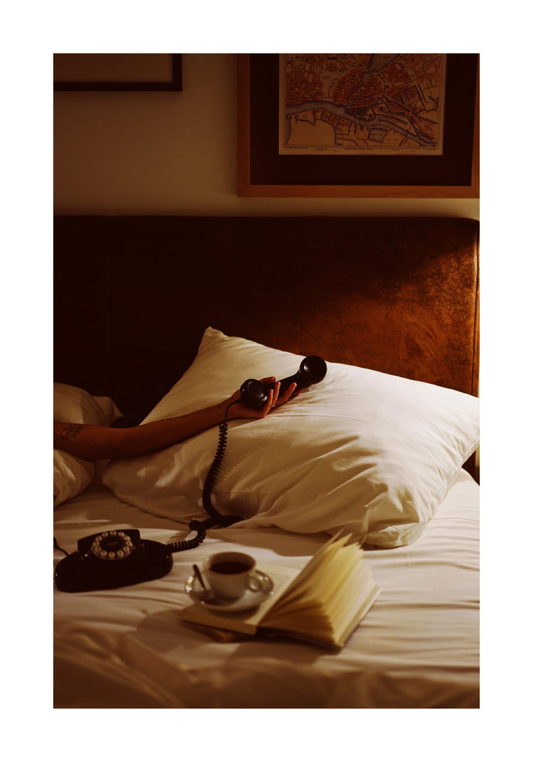 A person lies in bed, arm extended holding a vintage phone receiver. A white pillow and open book share the space, with coffee nearby. The Do Not Disturb Poster featuring a map design hangs above. Warm lighting enhances the cozy, retro vibe.