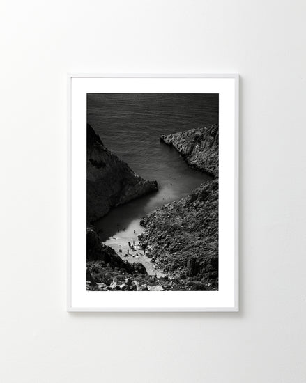 The A Crack in the Ocean poster, a framed black and white photograph of a rocky coastal inlet with a narrow beach, elegantly captures the sea extending beyond the cliffs. Displayed on a plain white wall, it exudes timeless elegance.