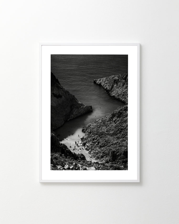 The A Crack in the Ocean poster, a framed black and white photograph of a rocky coastal inlet with a narrow beach, elegantly captures the sea extending beyond the cliffs. Displayed on a plain white wall, it exudes timeless elegance.