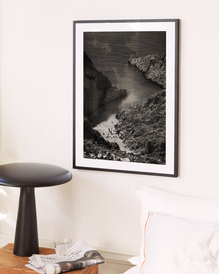 The A Crack in the Ocean Poster, a framed black-and-white photo of a rocky coast, hangs on a white wall above a bedside table with a glass of water, newspapers, and a minimalistic round lamp. A bed with white linens is partially visible, enhancing the rooms serene atmosphere.