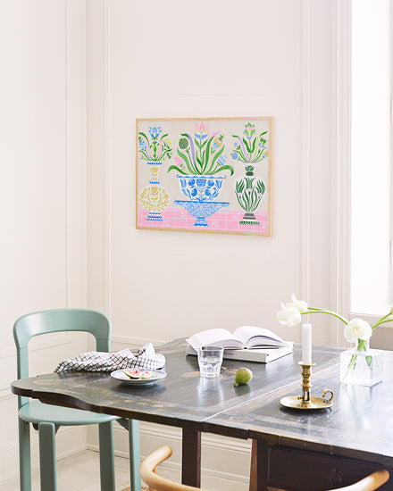 A dining table set against a light wall features the Ceramics Poster, with colorful framed botanical artwork reminiscent of Swedish kurbits. The table displays a green chair, checkered cloth, an open book, a glass of water, vibrant green fruit, and two white flowers in a brass candlestick holder.
