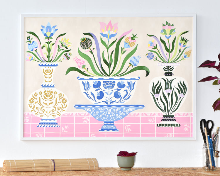 The Ceramics Poster features framed artwork of three decorative vases showcasing colorful geometric floral patterns inspired by Swedish kurbits. A blue central vase is flanked by vibrant vases set against a pink and beige background, with a bamboo mat and plant on the table.