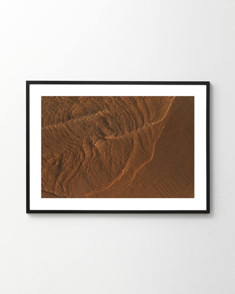 The Tide Poster, featuring brown sand patterns like moon-influenced tides, creates an abstract, earthy design with texture and shadows as it hangs on a white wall.