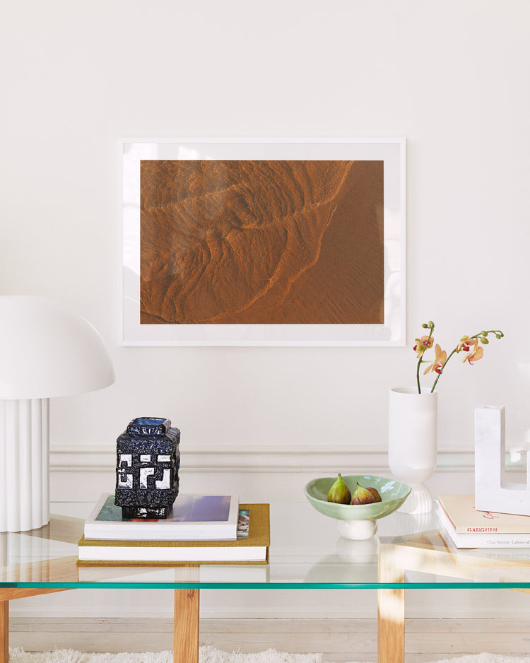 In a modern living room lit by moonlight, a glass coffee table holds books, a decorative blue vase, and green figs. A white lamp and an orchid adorn the table while the Tide Poster, depicting a desert landscape, hangs on the white wall.