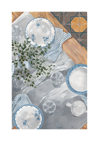 A tabletop set showcases The Good Plates Poster with blue and white floral patterns, features clear glasses, a mug, and is complemented by a striped cloth and leafy plant in a vase. The wooden surface harmonizes with the charming tiled floor.