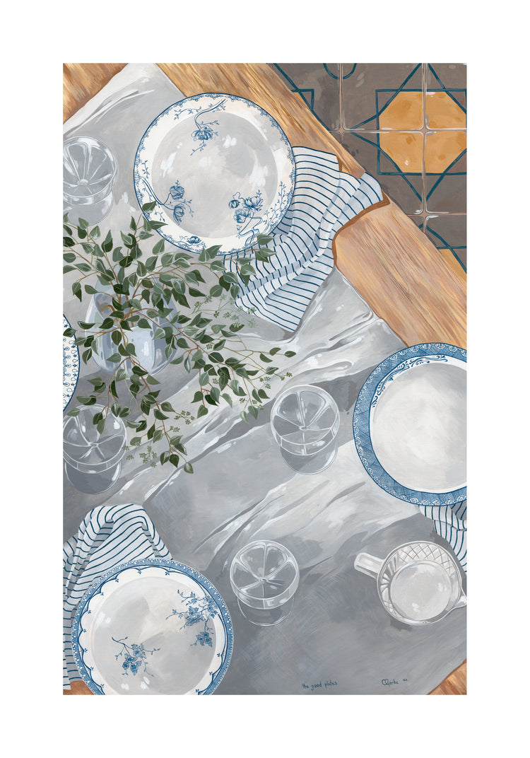 A tabletop set showcases The Good Plates Poster with blue and white floral patterns, features clear glasses, a mug, and is complemented by a striped cloth and leafy plant in a vase. The wooden surface harmonizes with the charming tiled floor.