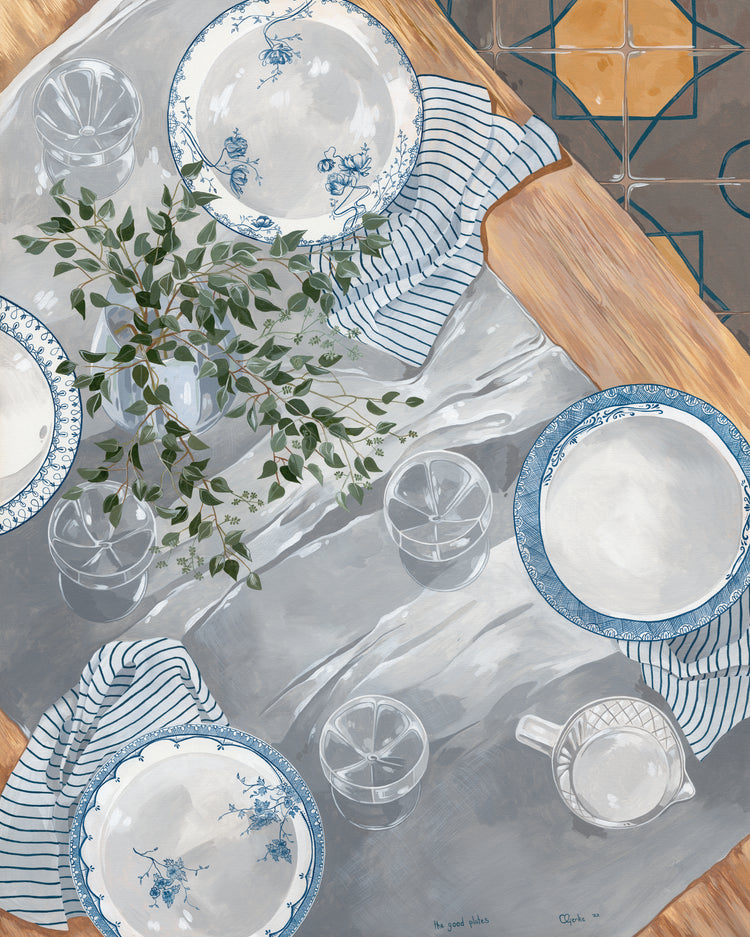 The Good Plates Poster features a dining table with decorative blue and white plates from the collection, paired with glasses and a striped cloth. A green leafy branch decorates the scene, while the floor displays brown and blue geometric tiles.