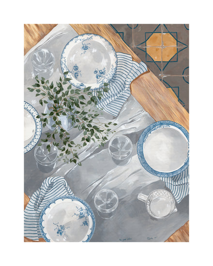 A table set with The Good Plates Poster in blue and white, glasses, a striped napkin, and a small pitcher on a wooden surface decorated with greenery. A tiled floor is visible in the corner.