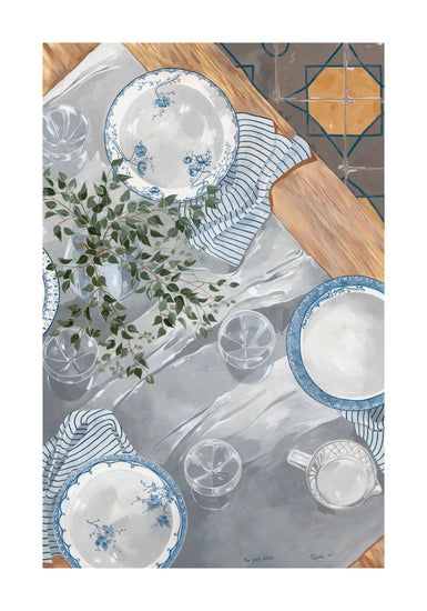 A table set with The Good Plates Poster flaunts blue and white florals on a shiny surface, paired with glasses, a striped cloth, and a small plant in the center. The wooden edge of the table complements the patterned floor visible in the background.