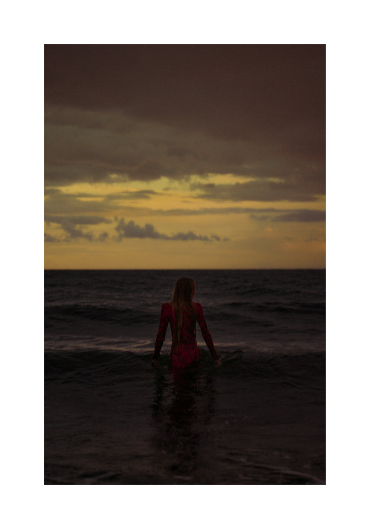 A woman in a red dressing gown walking into the ocean during sunset.