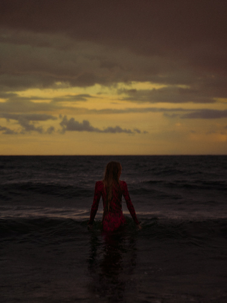 A woman in a red dressing gown walking into the ocean during sunset.