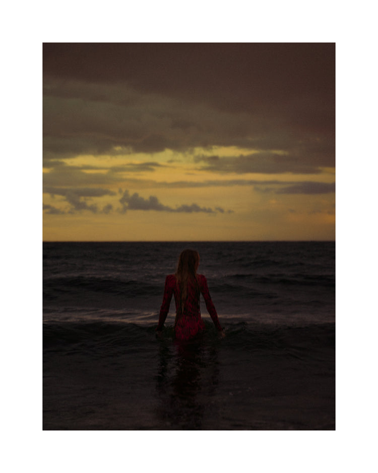A woman in a red dressing gown walking into the ocean during sunset.