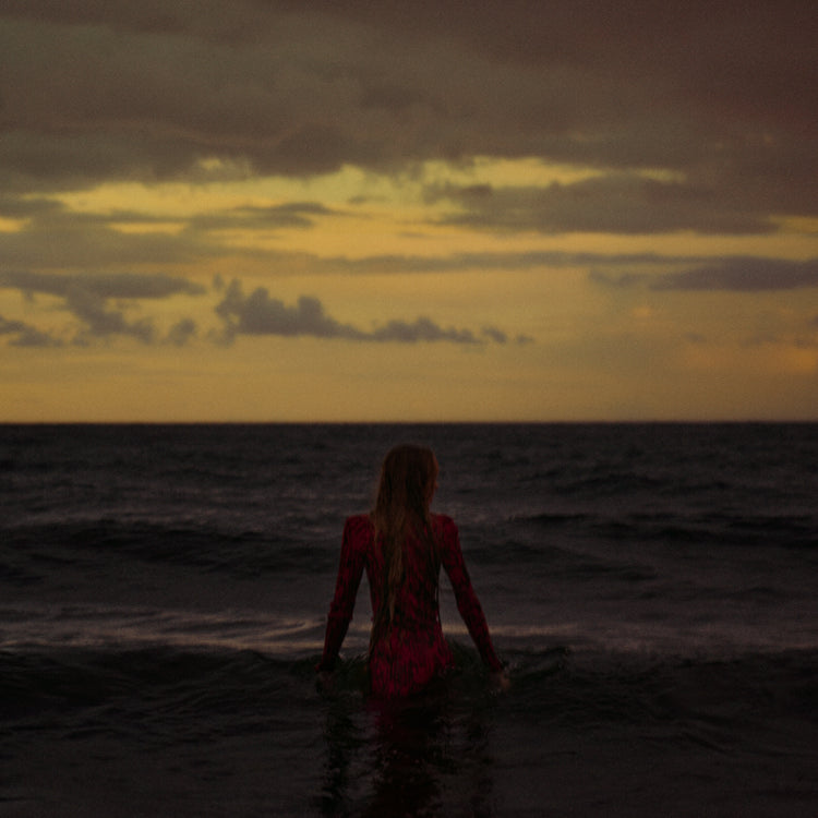 A woman in a red dressing gown walking into the ocean during sunset.