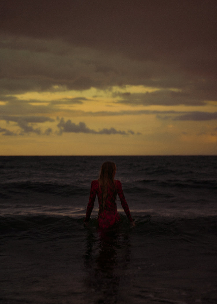 A woman in a red dressing gown walking into the ocean during sunset.