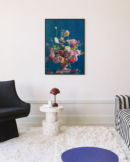 A modern minimalist living room showcases a black chair, terrazzo side table with a small red lamp, and blue circular table. An elegant white rug lays beneath the wall adorned with the colorful Pink Kiss Poster.